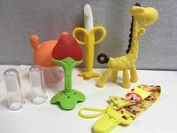 All seven baby teething items displayed: giraffe, banana toothbrush, strawberry teether, crab teether, two finger brushes and pacifier clip.