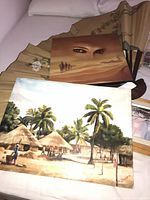 Both unframed paintings on bed with folding fan, showing village and desert scenes