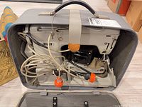 Sewing machine inside open gray case with cords and foot pedal visible
