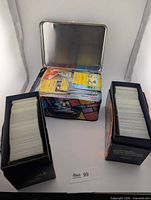 Two Elite Trainer Box containers filled with stacked cards alongside open metal lunch tin showing additional stacks
