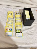 Rows of yellow-border Pokémon Trainer/Item/Supporter cards laid out beside black storage deck box