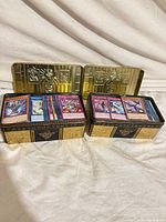 Two gold Yu-Gi-Oh! tins open showing stacks of assorted cards
