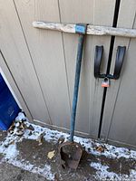 Full view of post hole digger leaning against shed showing handle, shaft and scoop head
