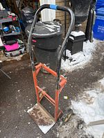 Full view of dolly upright on driveway with snow, showing handle, frame, wheels and platform
