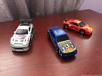 Three Hot Wheels cars on wooden surface: white Supra, blue Stocker, red F355