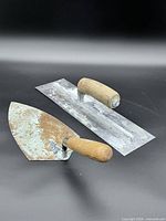 Rectangular finishing trowel and triangular masonry trowel with wooden handles displayed on black background