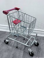 Front angle of metal toddler shopping cart showing red handle and flip-up seat