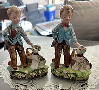 Two identical Arnart Creation Japan porcelain figurines of schoolboys with terrier dogs on grassy bases.