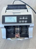 Front view of IdleTech BC-1100 money counter showing display, controls, bill path and attached power cord