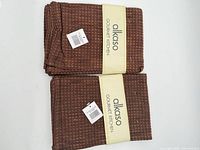 Two factory-folded stacks of chocolate brown waffle tea towels with Alkaso bands