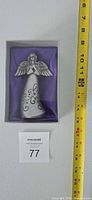 Front view of boxed silver-tone angel figurine beside measuring tape showing roughly 5 inch height