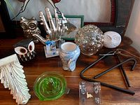 Assorted decor laid on tabletop including metal hand sculpture, uranium glass juicer, owls, dinner bell pieces and more