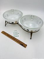 Two casserole dishes with lids on metal stand next to ruler