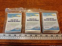 Three packaged Primacare foil mylar blankets side by side with ruler for scale