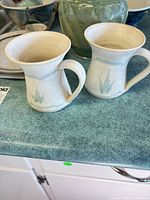 Two cream stoneware mugs; partial carved green vase and bowls visible