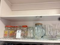 Shelf view showing six floral-decal jars with lids, pressed glass compote, wire-bail jar, aqua cork-top jar, mug, sugar bowl, small pitcher