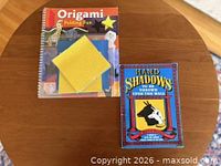 Front covers of Origami Folding Fun spiral book with yellow origami papers on top and Hand Shadows paperback by Henry Bursill