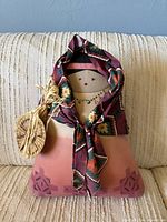 Front view of stuffed cloth doll showing painted face, patterned headscarf, raffia and woven basket accent