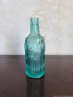 Front view of teal pressed glass bottle showing ribbed body and floral embossing