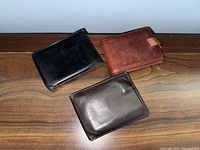 Three leather wallets on wooden surface: black, reddish-brown with tab, dark brown