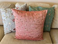 Front view of three embroidered cushion covers in pink, light blue-grey and green