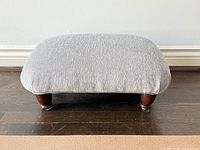 Front view of grey upholstered footstool showing fabric, shape and wooden legs