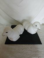 Both white ceramic lamb figurines side by side on black mat