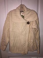 Front view of cream merino wool cardigan showing shawl collar, cable patterns, button, hang tag