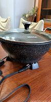 Electric wok on table showing speckled body, glass lid, power cord attached