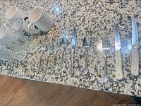 Flatware, steak knives, ceramic and glass mugs laid out on countertop