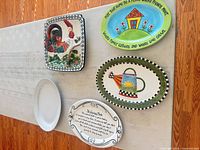 Five ceramic plates displayed on table: rooster platter, house scene plate, watering can platter, giving plate, plain white plate