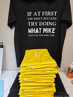 Stack of sealed yellow poly bags with one black T-shirt displayed showing full text
