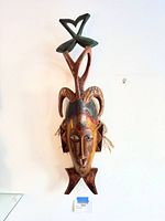 Full view of carved wooden tribal mask on wall showing horns, finial, tassels, and finish