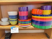 Full lot showing stacks of multicolor plastic plates, plastic bowls, ceramic bowls, saucers and teacup on shelf