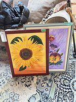 Front view of two framed oil paintings (sunflower and pansy) leaning on rug