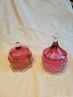 Both cranberry glass candy dishes side by side with lids on