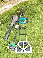 All items laid out on grass: gas leaf blower, gas string trimmer, folding hand truck, fish tape reel, hedge shears