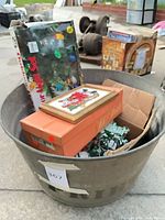 Metal tub holding boxed outdoor lights, cardboard box, framed Santa needlepoint and other decorations