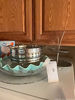 All four kitchenware items grouped together: ruffled glass dish atop glass platter, stainless colander stacked inside, two-tier stand beside