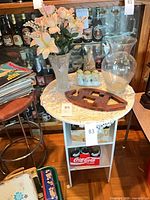 Overview showing both glass vases with flowers, porcelain figures, wooden trivet on round table, and Coca-Cola bottles below