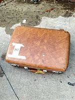 Closed brown hard-shell suitcase on pavement, showing exterior condition and latch placement