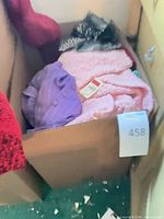 Box containing assorted garments including pink fleece, purple fabric, and patterned piece