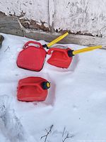 Three red plastic fuel cans lying in snow beside wall; two with yellow spouts