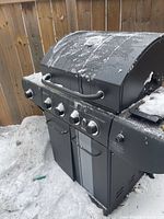 Front view of snow-covered black propane grill showing four control knobs, lid thermometer, cabinet doors