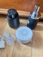 Sound machine, USB adapter with cable, matte black soap dispenser, matte black lidded container grouped on wood floor
