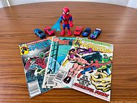 Group shot of all three comics, action figure and five toy cars on tabletop