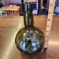 Full view of olive green demijohn bottle beside ruler showing ~12 in height