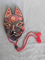 Front view of black kitsune mask showing painted red and gold designs and tasselled cord