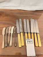 All twelve butter utensils laid out: five tarnished silver-plated spreaders and seven yellow-handled stainless knives