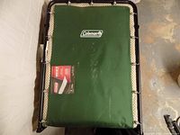 Top view of folded Coleman cot showing green fabric deck, branded logo, beige patterned pad and springs
